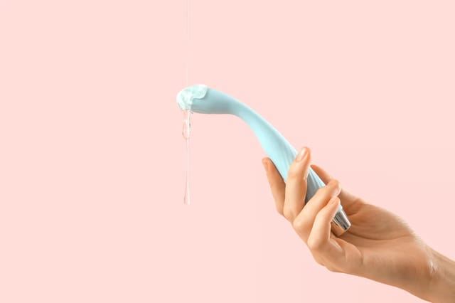 blue-curved-intimate-massager-with-gel Hand holding a blue curved intimate massager with clear gel against a soft pink background.