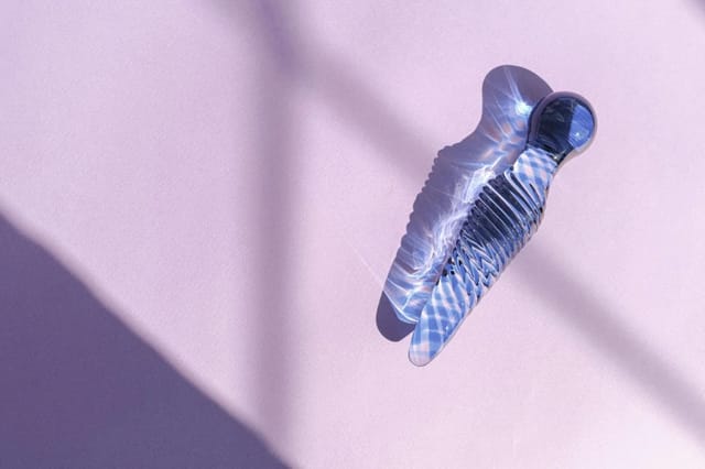 blue-glass-intimate-toy-on-lavender-background Blue transparent glass intimate toy on a lavender surface with soft sunlight and shadow patterns.
