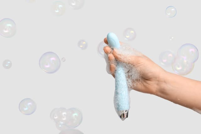 blue-waterproof-vibrator-cleaning-with-bubbles Hand washing a blue waterproof vibrator with soap bubbles against a clean white background.