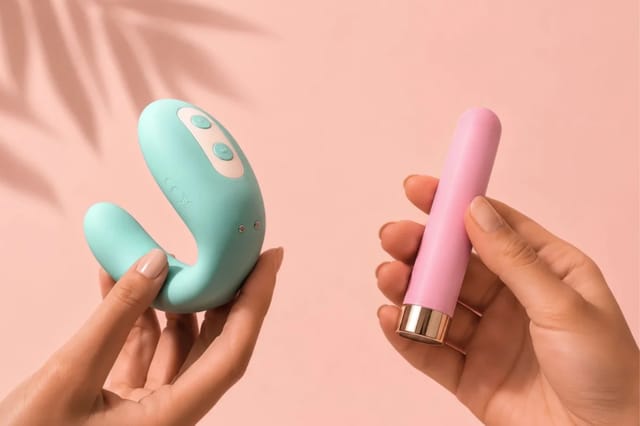 hands-holding-blue-vibrator-and-pink-bullet-vibrator Hands holding a teal clitoral vibrator and a pink bullet vibrator against a soft peach background.