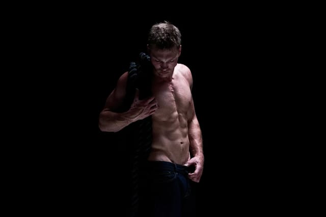 male-body-moody-studio-lifestyle Moody studio lifestyle image of a shirtless adult man holding fabric against a black background.