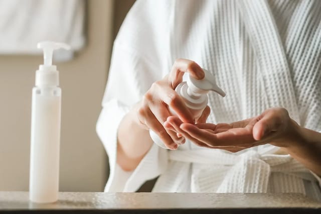 woman-applying-body-lotion-in-white-robe Woman in a white robe dispensing body lotion into her hand in a soft bathroom setting.